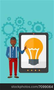 An african-american man pointing at a big tablet computer with a light bulb on a screen on a blue background with cogwheels vector flat design illustration. Vertical layout.. Man pointing at tablet computer with light bulb on screen.