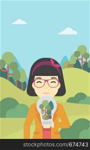 An asian woman standing on the background of green forest and holding in hands a big lightbulb with trees inside. Green energy concept. Vector flat design illustration. Vertical layout.. Woman with lightbulb and trees inside.