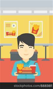 An asian young man holding tray full of junk food on a cafe background. Smiling man in fast food restaurant. Man having lunch in a fast food restaurant. Vector flat design illustration. Vertical layout.. Man with tray full of fast food.