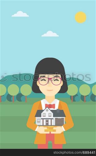 An asian young woman holding house model in hands on the background of mountains. Real estate agent with house model in hands. Vector flat design illustration. Vertical layout.. Woman holding house model vector illustration.