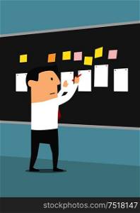 Businessman pinning sheets of paper and stickers on blackboard with concentrated or serious facial expression. Manager making business plan or reaching goal, setting task or posing problem. . Businessman pinning sheets of paper on blackboard