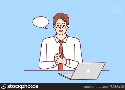 Confident businessman is sitting at table with laptop and looking at camera recording business training. Guy in business clothes with dialogue cloud works as teacher online training courses . Businessman is sitting at table with laptop and looking at camera recording business training