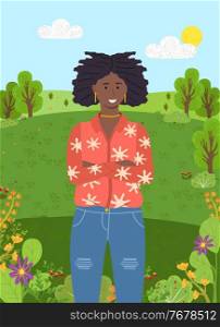 Dark-skinned confident young smiling girl with dreadlocks and gold jewelry stands with her arms crossed on her chest. Fashionable ripped jeans, bright jacket. Green city park or forest on background. Dark-skinned young girl stands on background of green forest or park. Trees, clear sky, sun