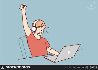 Delighted teen boy sitting at table with laptop playing video game and defeating virtual opponents. Schoolboy in headphones makes victory gesture overjoyed at broken record in internet game . Delighted teen boy sitting at table with laptop playing video game and defeating virtual opponents