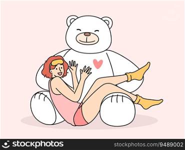 Giant teddy bear near sitting teenage girl in pajamas and sleep mask. Funny schoolgirl teen age looks at screen lying on feet of toy bear rejoicing at cute gift from parents or boyfriend. Giant teddy bear near sitting teenage girl in pajamas and sleep mask rejoicing at cute present