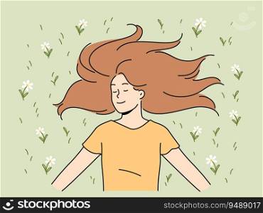 Happy woman with long hair lies on grass in park with eyes closed and smiles rejoicing in sunlight. Positive young woman in casual t-shirt enjoying good summer weather lying on lawn with flowers. Happy woman with long hair lies on grass in park with eyes closed and smiles rejoicing in sunlight