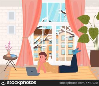 Leisure time at home, man lying on carpet and watching film at laptop, snowy winter outside of window, snowman, relaxing indoors, stylish interior with houseplant and table with decor, young guy rest. Leisure time at home, man lying on carpet and watching film at laptop, snowy winter outside
