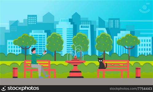 Man sitting on the park bench and holding a small bird in his hand a sad black cat sits nearby. Male walking in a city park with benches a fountain and an alley against the backdrop of the cityscape. Man sitting on the park bench and holding a small bird in his hand a sad black cat sits nearby