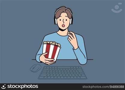 Man with package of popcorn watches movie sitting at table with computer and looks at screen intrigued. Funny guy in headphones is relaxing watching new movie blockbuster or popular tv series. Man with popcorn watches movie sitting at table with computer and looks at screen intrigued