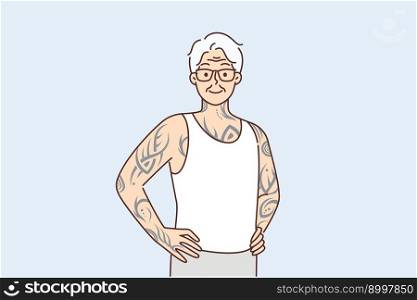 Optimistic elderly man with tattoos on arms and body stands with hands on belt proud good health in old age. Gray-haired elderly human with tattoos stuffed in youth looks at screen posing in t-shirt. Elderly man with tattoos on arms and body stands with hands on belt proud good health in old age