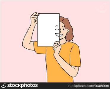 Smiling woman holding piece of paper with emoji in front of face and looking at screen. Positive girl with partially painted smile is sharing good mood and positive emotions with you.. Smiling woman holding piece of paper with emoji in front of face and looking at screen