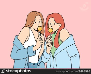 Two women with chupa chups in hands dressed in fashionable seductive clothes eat lollipops. Lovely girls in stylish jackets slung off shoulders to advertise new candies for teenagers. Two women with chupa chups in hands dressed in fashionable seductive clothes eat lollipops