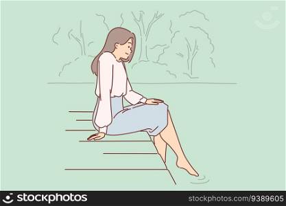 Woman is sitting on pier, lowering into water and enjoying silence and good summer weather. Dreamy young girl looks at river or lake relaxing on pier in public park after hard day work.. Woman is sitting on pier, lowering into water and enjoying silence and good summer weather