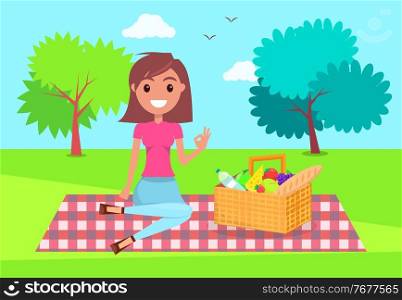 Young girl sitting at checkered plaid in park with basket full of products. Smiling girl relaxing at nature, summer time. Pretty female wearing t-shirt, jeans resting at picnic. Girl show ok sign. Young brunette girl at picnic in summer park, lady sitting at plaid with basket full of products